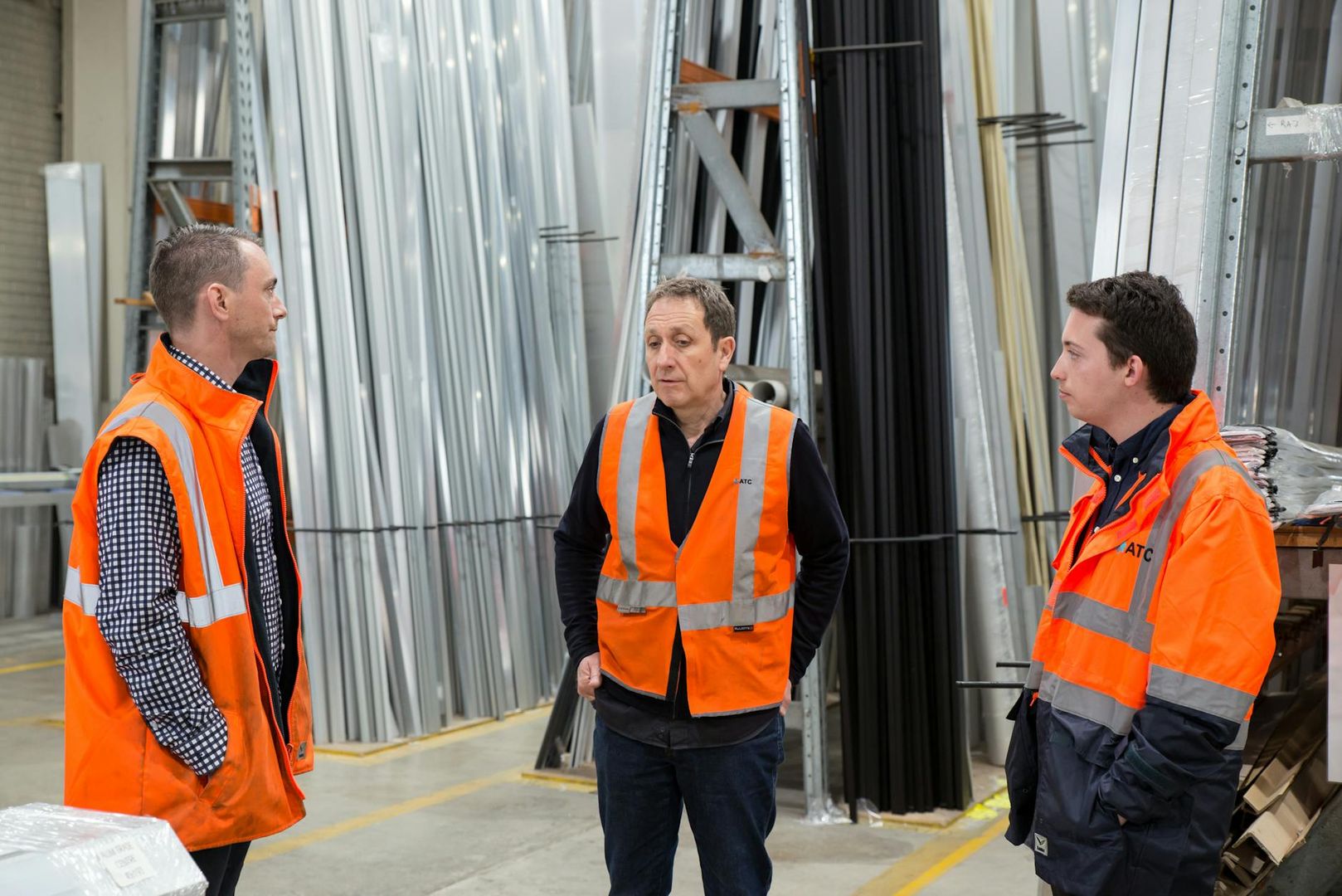 Tre lagerarbetare i orangea varselvästar diskuterar i ett lager för metallprofiler.